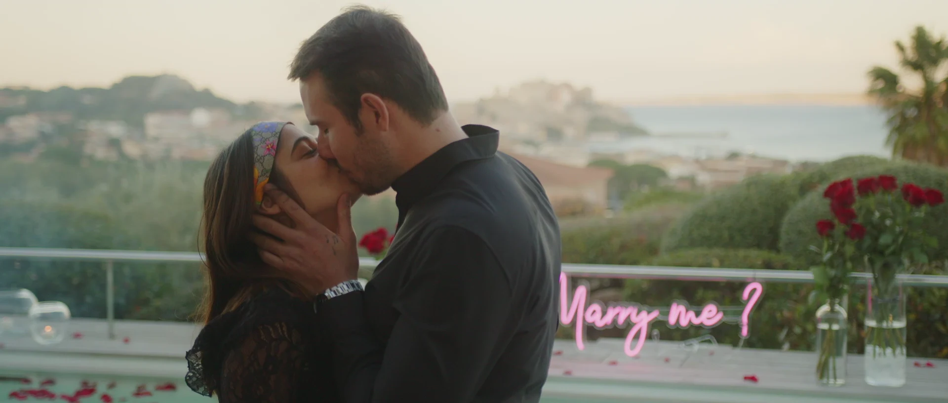 Couple kissing just after the proposal, 'Marry me?' neon sign behind them